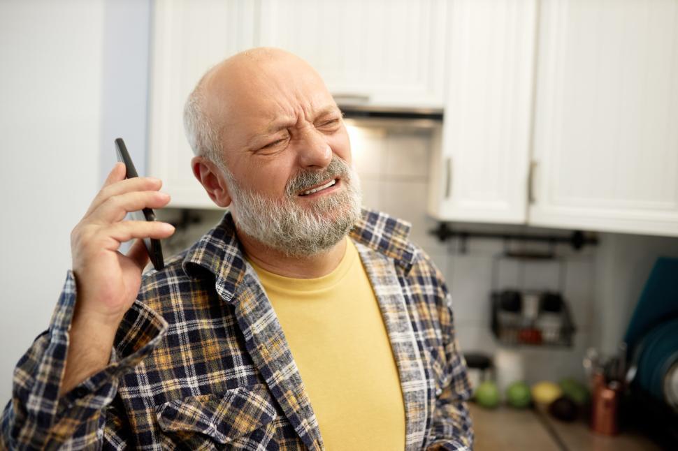 Free Image of Squinting displeased mature handsome husband listening to voice mail or having phone conversation with his furious angry wife yelling at him through smartphone, keeping it far from his ear 