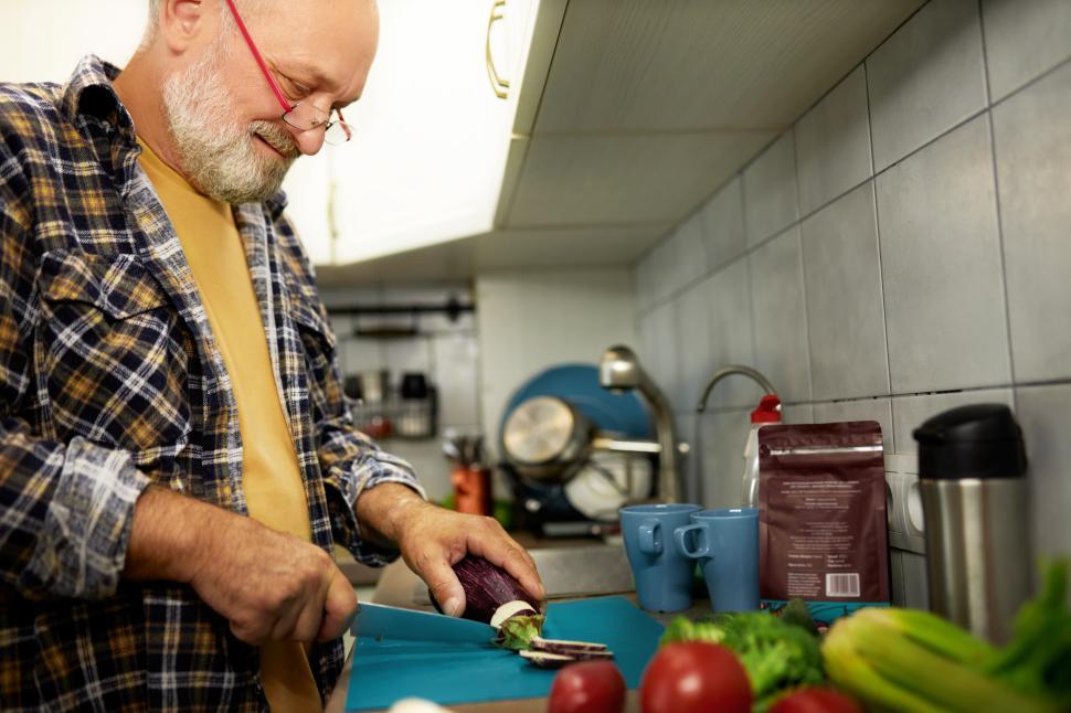 Free Image of Side low angle view of senior happy man chopping and cutting organic vegetables with knife on counter in kitchen to cook delicious healthy dinner. Grandpa preparing breakfast for his family 