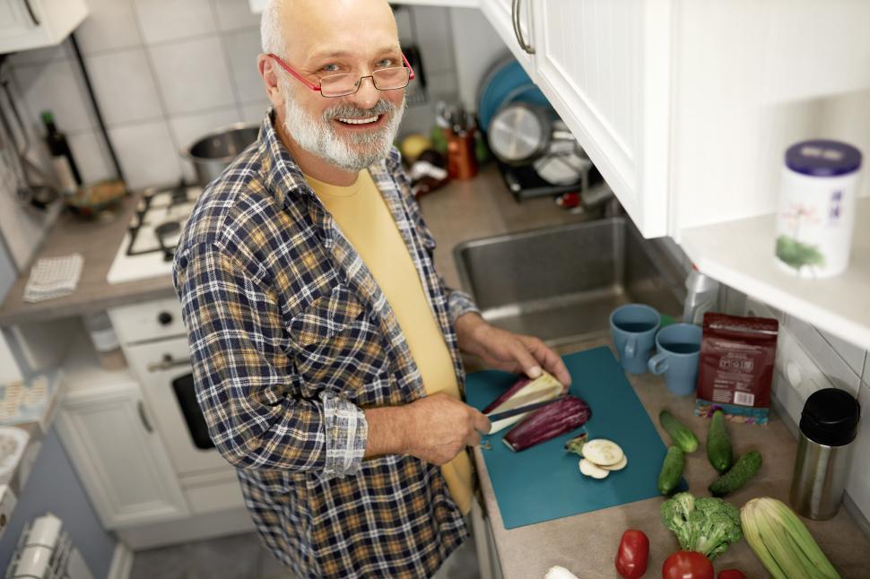 Free Image of Upper view of happy mature husband of father chopping aubergine to cook breakfast for his family, looking at camera with happy smile, holding sharp knife, standing in kitchen on sunday morning 