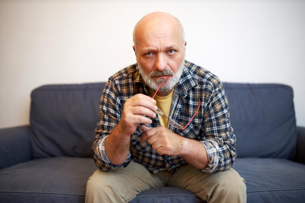 Free Image of Pensive thoughtful senior bald bearded man sitting on couch biting ends of his glasses looking at camera with contemplation, trying to find answer on philosophic question and find sense of life 
