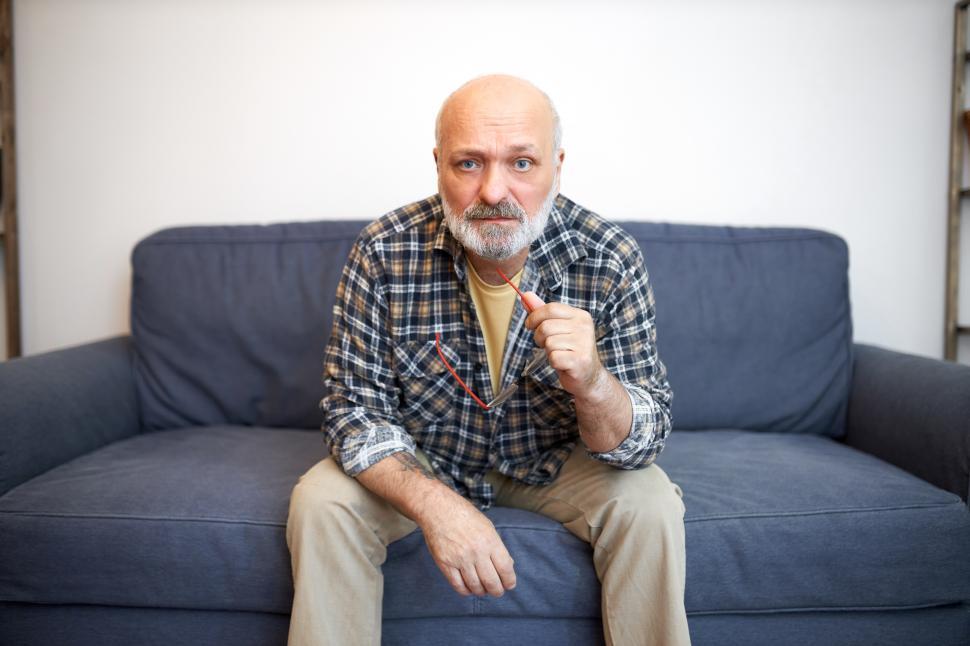 Free Image of Indoor portrait of worried anxious grandfather in plaid shirt having rest sitting on couch in living room holding glasses, thinking of his life, world problems, health and aging process 