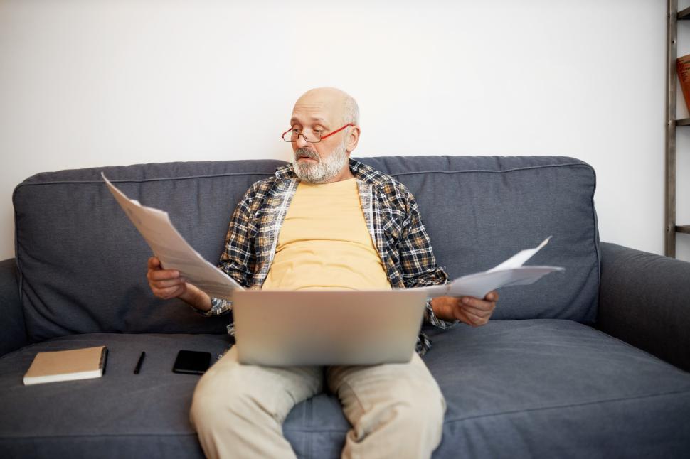 Free Image of Portrait of upset confused grandfather holding paper documents trying to figure out how to pay bills and loan, sitting on couch with laptop on knees, wearing glasses, reading agreement 