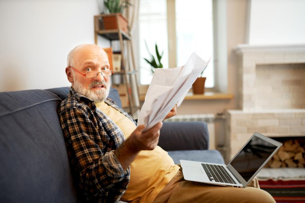 Free Image of Side view of worried anxious elderly man in glasses looking at camera with panic facial expression holding overdue bills, trying to do online payment using laptop with copy space screen 