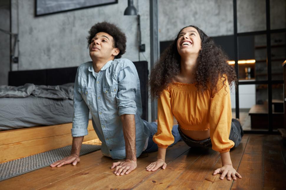 Free Image of Side view portrait of funny man and woman of 20s pretending to practice yoga on floor in casual clothes, standing in upward facing dog position in bedroom, before going out on sunday 