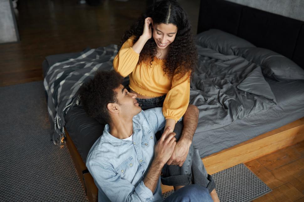 Free Image of Upper view portrait of happy afro guy in denim shirt sitting on floor next to his girlfriend resting on bed, touching her legs, showing his affection, love and loyalty. Human relationships 