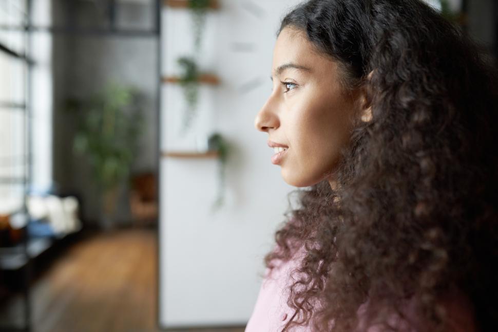 Free Image of Profile indoor view of gorgeous african american young female with clear skin and beautiful long curly hair looking ahead standing in stylish living room with pensive face. Copy space of the right 