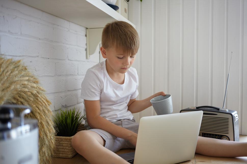 Free Image of Handsome 10 year old Caucasian boy in casual clothes sitting on kitchen counter, drinking tea and watching cartoons online using high speed wireless internet connection on generic laptop computer 