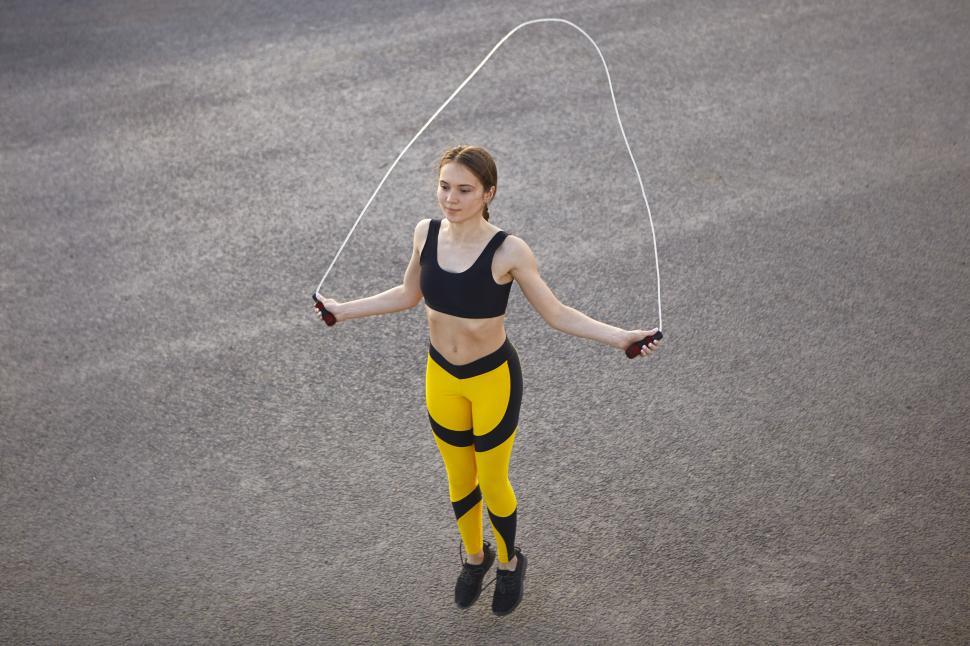 Free Image of Isolated full length shot of self determined young athletic woman in black and yellow sports outfit exercising outdoors with jumping rope against blank asphalt background with copy space for your text 