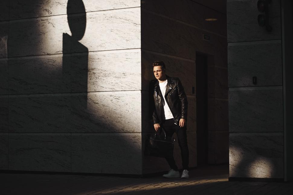Free Image of Full length shot of fashionable young European man wearing black jeans, leather jacket, white sneakers and t-shirt holding bag and leaning against modern building, waiting for his cab or colleague 