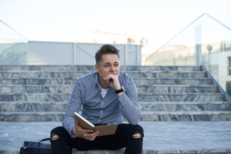 Free Image of Outdoor image of thoughtful smoothly shaved young Caucasian businessman in elegant stylish clothes sitting on concrete stairs in cityscape, holding open copybook, having pensive look, hand on his chin 