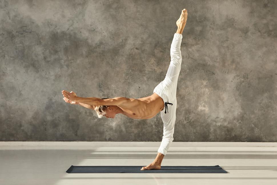 Free Image of Sideways indoor shot of sporty muscular man with naked torso doing Urdhva Prasarita Eka Padasana on mat, stretching and strengthening muscles, bending forward in parallel with floor, legs in split 