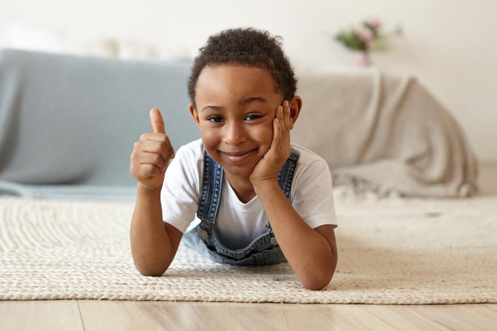 Free Image of Indoor image of funny adorable African American little boy in casual clothes lying on his stomach on floor at home and making thumbs up gesture, showing positive reaction, expressing approval 