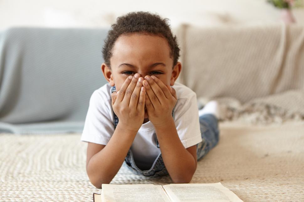 Free Image of Cheerful little Afro American boy lying on stomach on carpet in bedroom, covering mouth as if laughing, reading book or doing home assignment. People, childhood, learing and education concept 