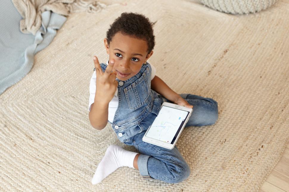 Free Image of Indoor image of adorable dark skinned black male kid wearing white socks and denim jumpsuit sitting on floor with generic digital tablet on his knees, looking at camera, showing victory sign 