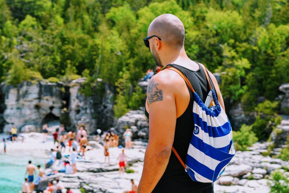 Free Image of Man with striped backpack by rocky lakeshore on sunny day. 