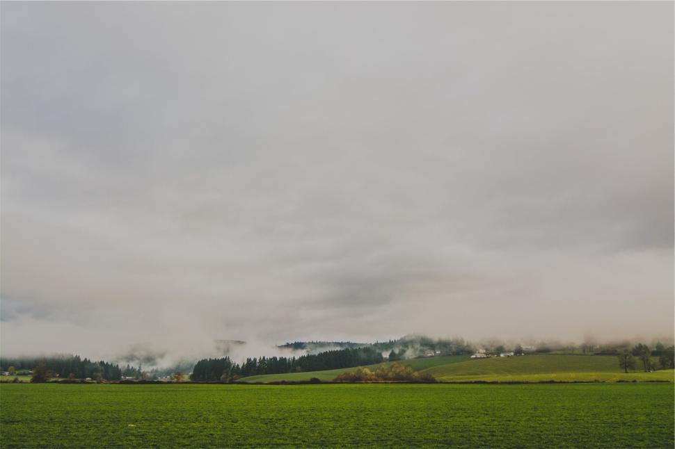 Free Image of Misty green field with hills and clouds in background 