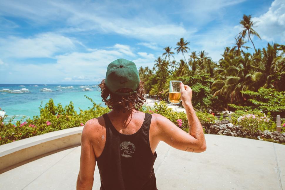Free Image of Man enjoying a beer facing the ocean and palm trees 