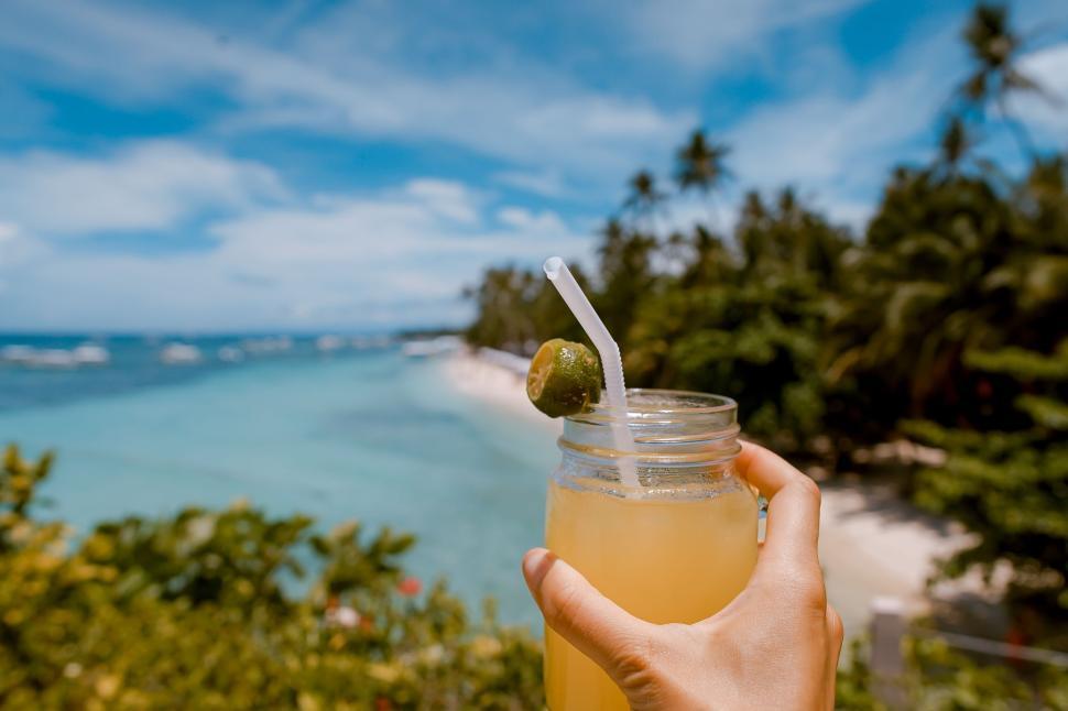 Free Image of Hand holding lemonade jar with straw on a beach day. 