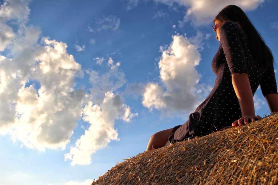 Free Image of Woman relaxing on haystack under a sky with fluffy clouds. 