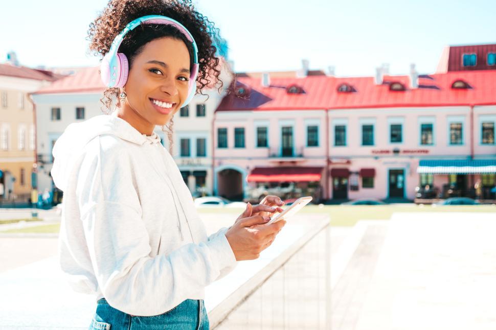 Free Image of Woman with headphones smiling, holding smartphone outside. 