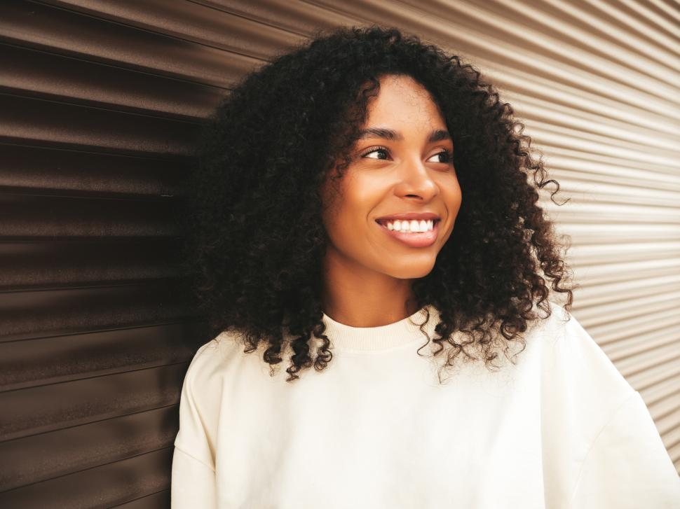 Free Image of Woman with curly hair smiling, standing against brown wall. 