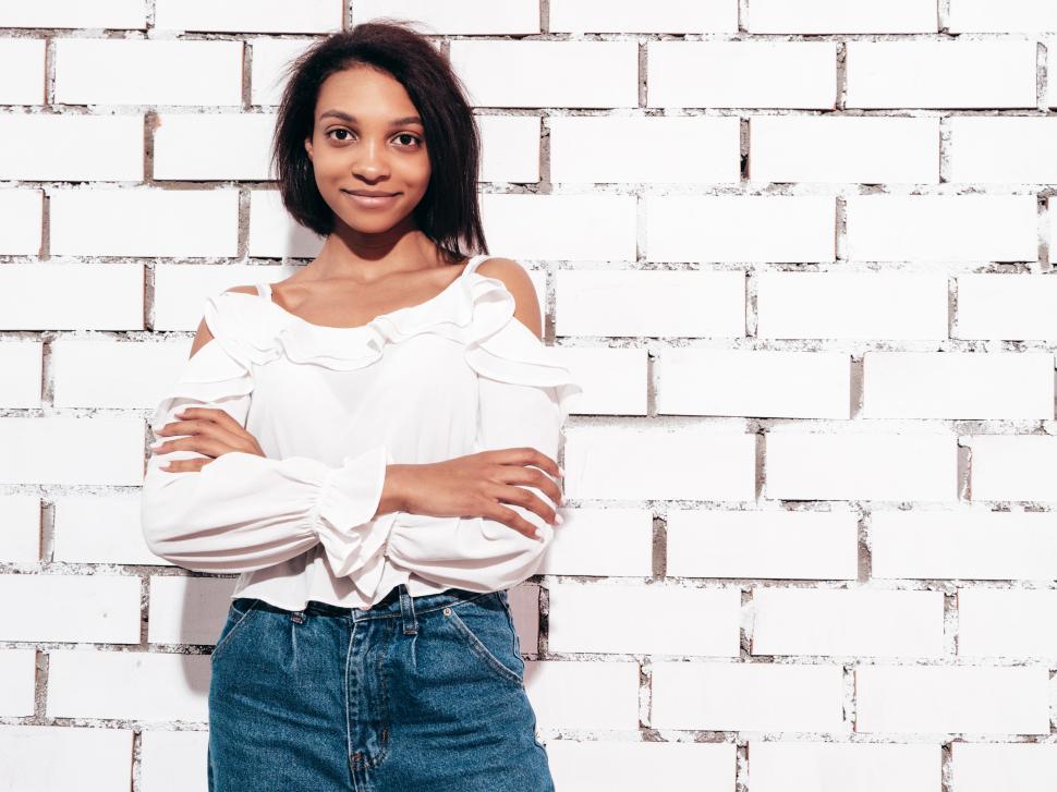 Free Image of Woman in white top and jeans posing against brick wall. 