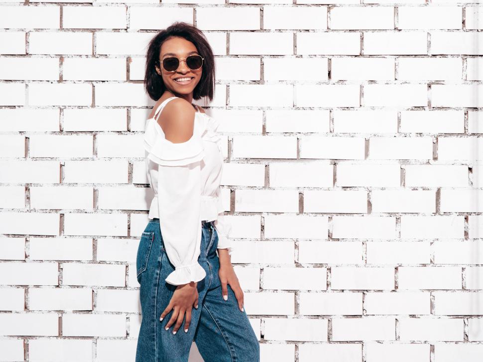 Free Image of Woman in white blouse and jeans smiling by brick wall. 