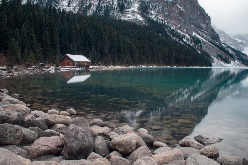 Free Image of Serene mountain cabin by clear lake with rocky shore. 