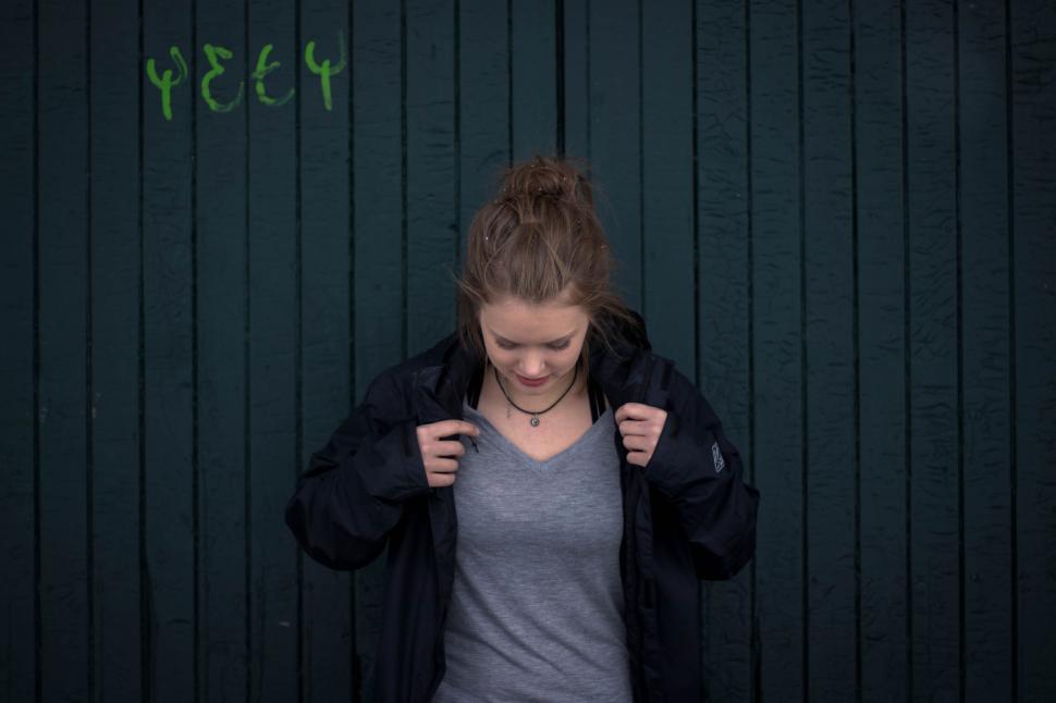 Free Image of Young woman in black jacket stands against green wall. 