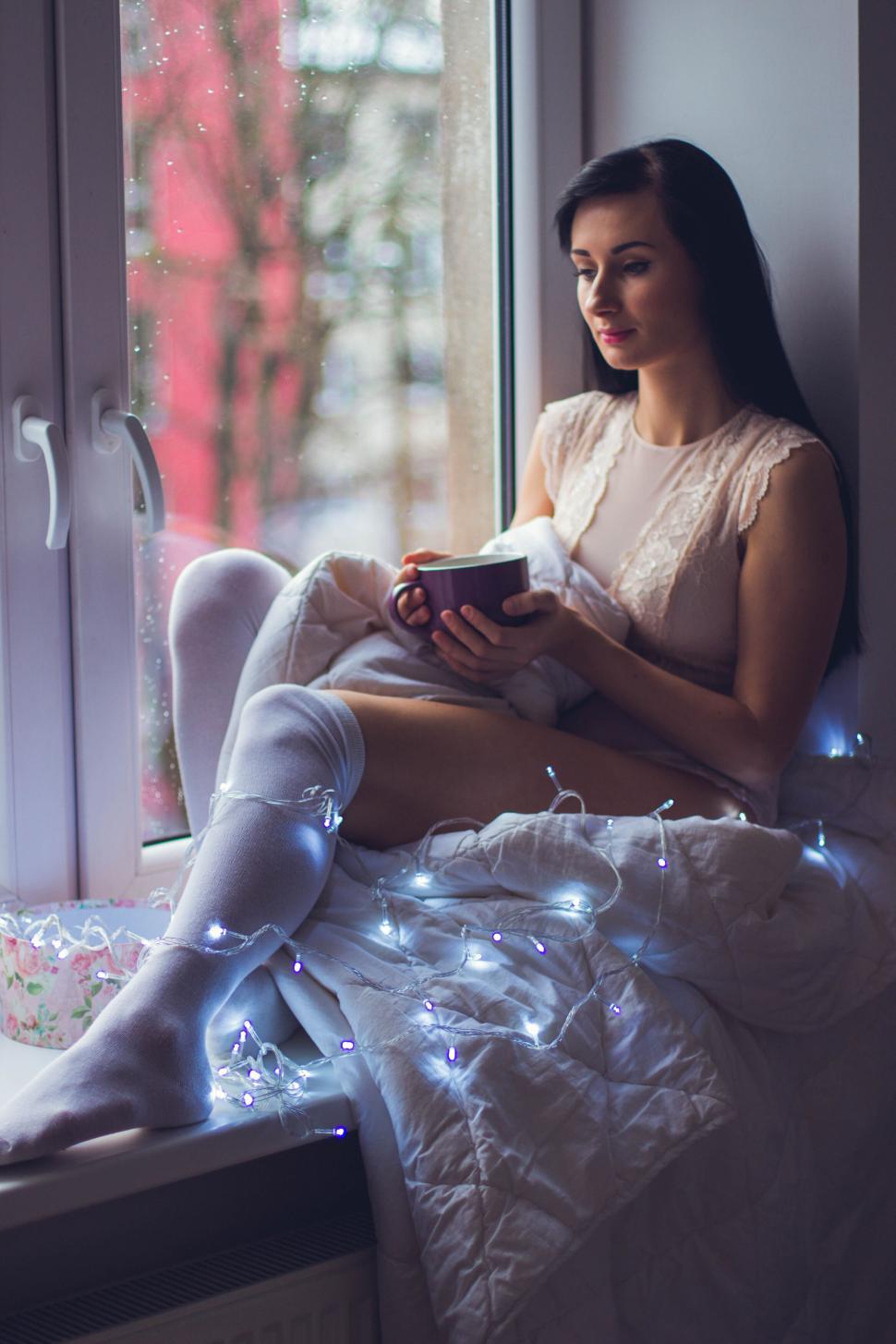 Free Image of Woman sitting by window with cup and fairy lights in lap 
