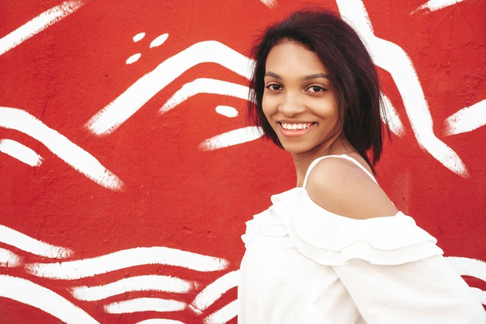 Free Image of Woman smiling, red wall, white off-shoulder top, casual jeans 