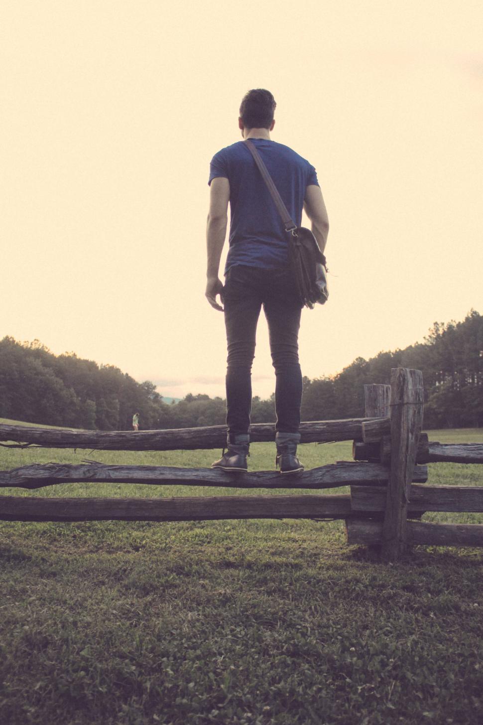 Free Image of Person standing atop a fence, bag slung over shoulder. 