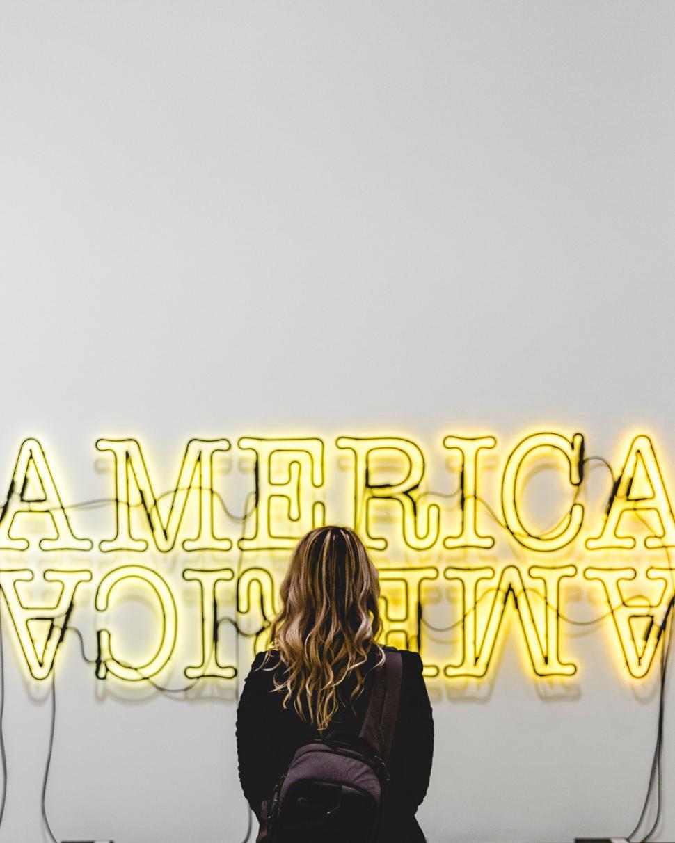 Free Image of Person with blonde hair stands in front of neon America sign 