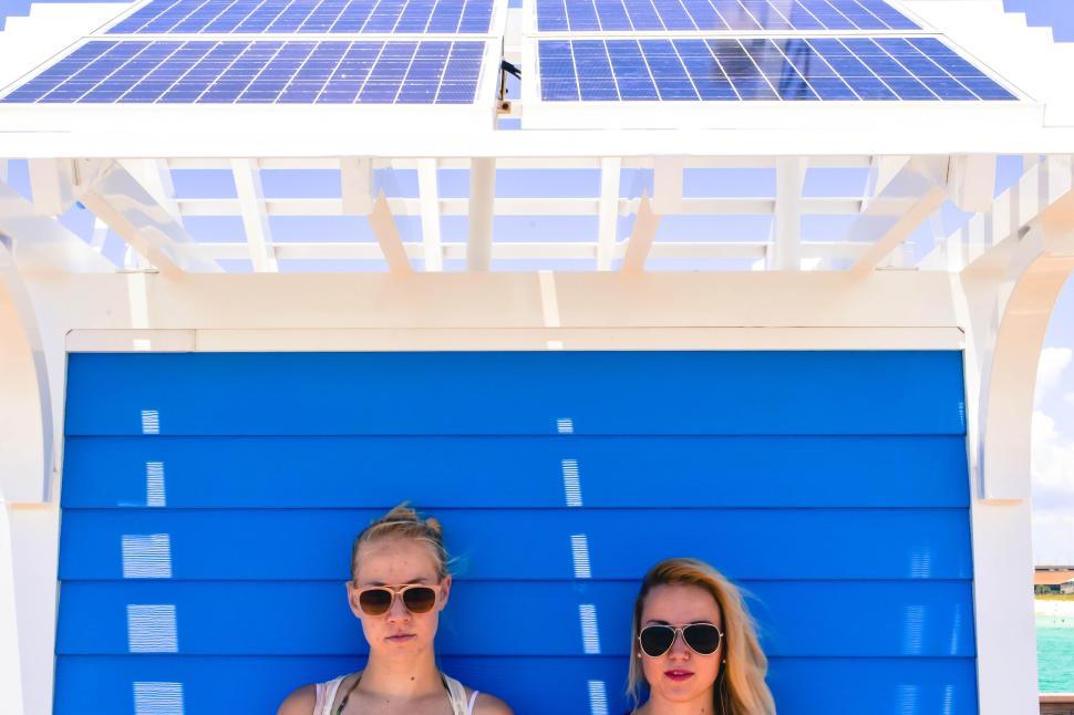 Free Image of Two women in sunglasses under solar panels on a beach. 