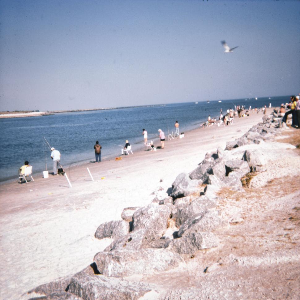 Free Image of Beachside with people relaxing and fishing near rocks. 