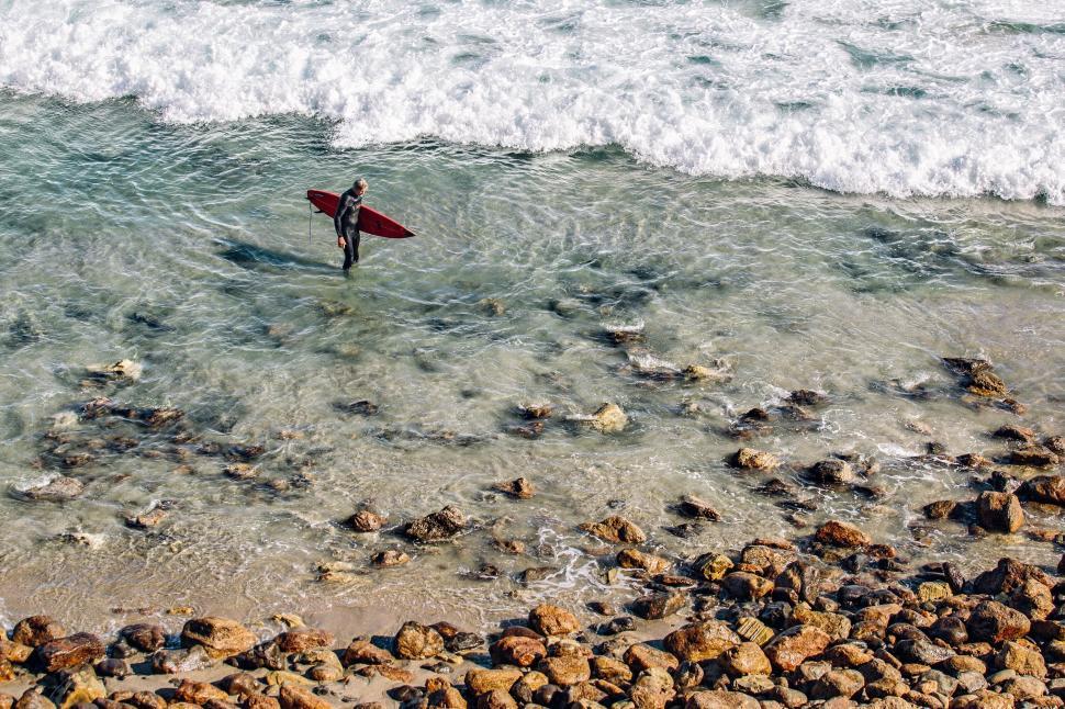 Free Image of A surfer with a red board stands on a rocky shoreline. 