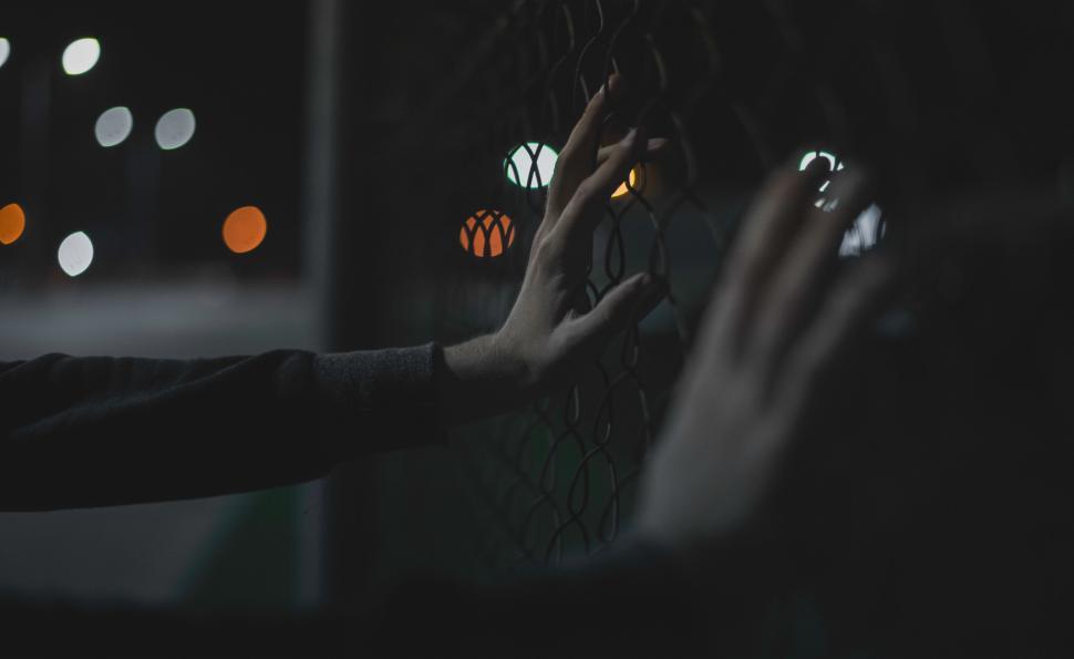 Free Image of Hands holding a fence at night with blurred lights behind. 