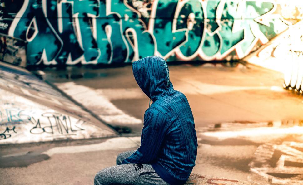 Free Image of Person in a blue hooded jacket sitting by graffiti wall. 