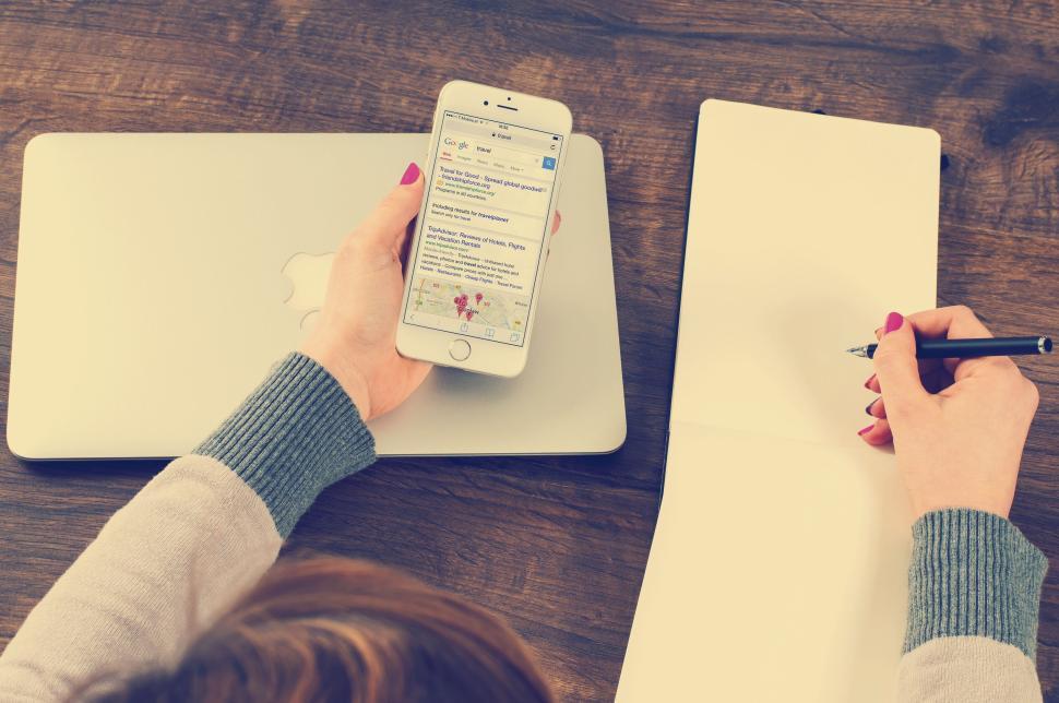 Free Image of Person holding phone, writing in notebook, laptop nearby. 
