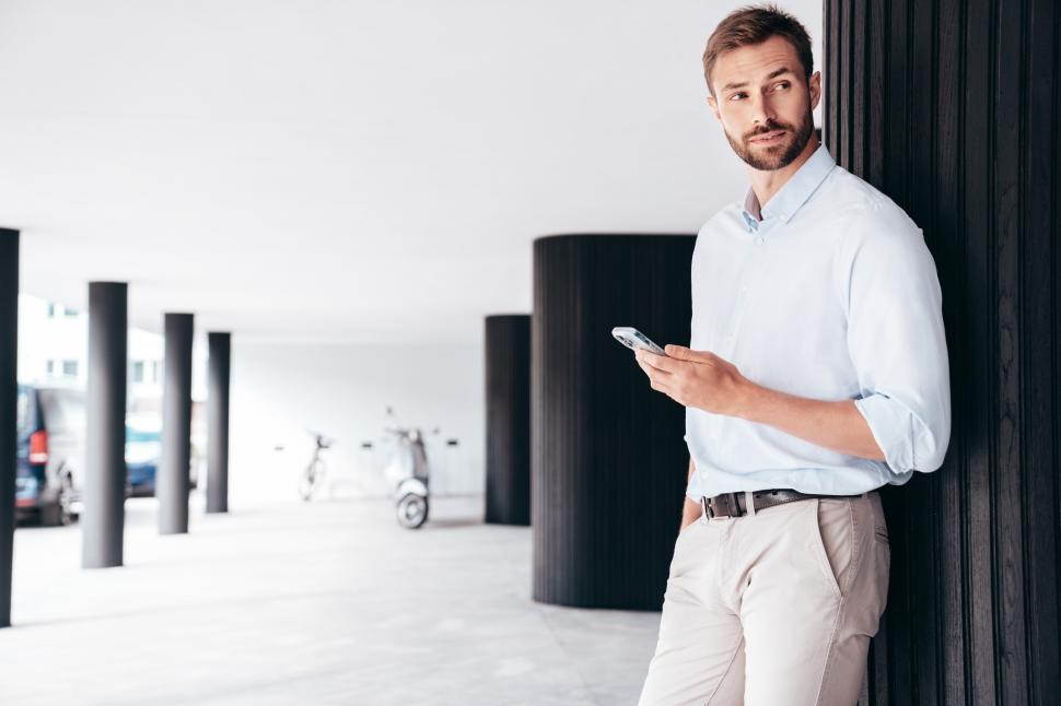 Free Image of Young man using smartphone while leaning against a pillar in modern urban setting during daytime 