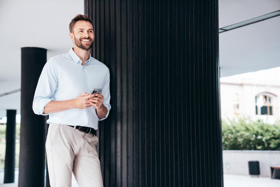 Free Image of Man leaning against a column while using a smartphone in an urban setting with modern architecture 