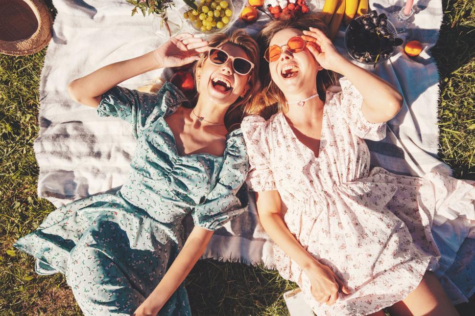 Free Image of Two friends enjoy a sunny picnic with fresh fruit in a park while wearing stylish summer dresses and sunglasses 