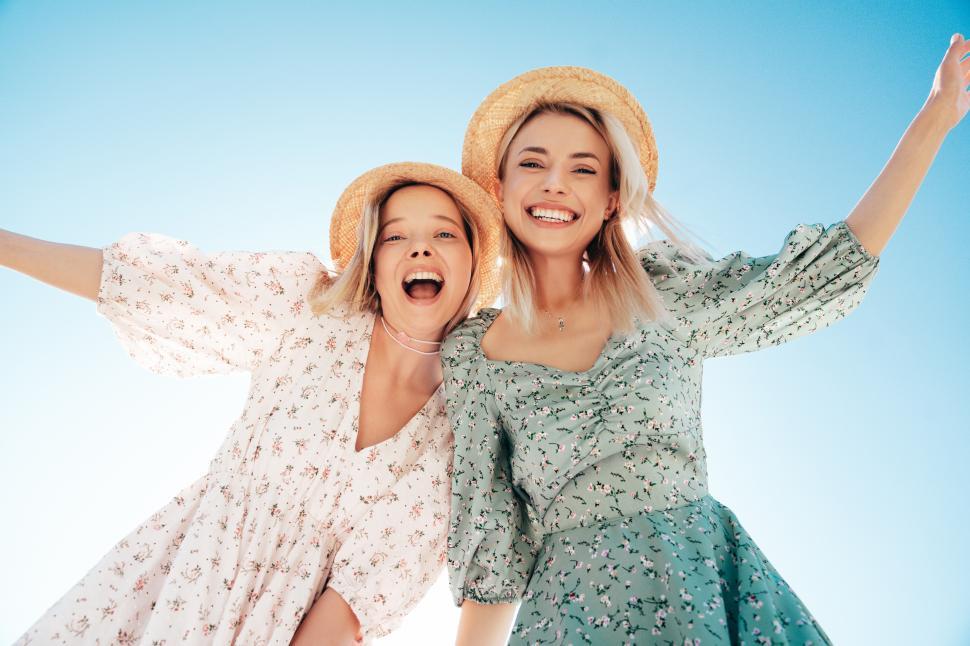 Free Image of Two young women joyfully posing under a blue sky wearing summer dresses and straw hats while enjoying a sunny day outdoors 
