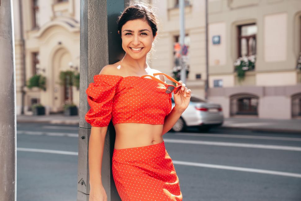 Free Image of Young woman wearing a red polka dot outfit poses confidently on a city street during sunny weather 