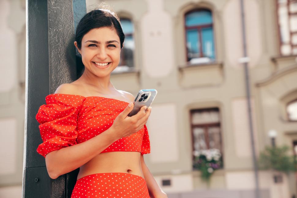 Free Image of Smiling woman in red polka dot outfit enjoys a sunny day while using her smartphone in a city street 