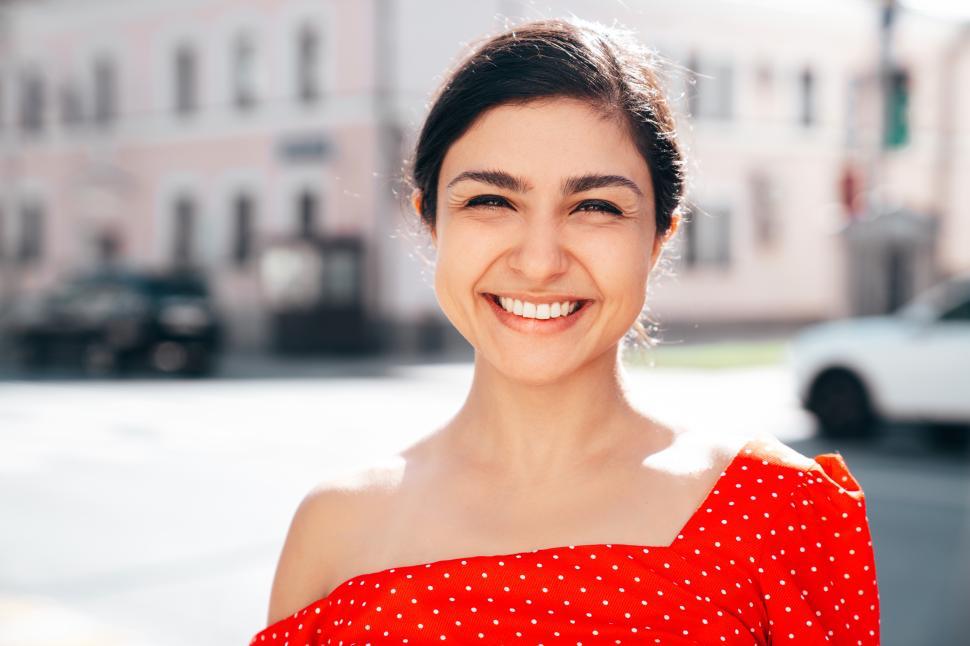 Free Image of Joyful young woman smiles brightly outdoors wearing a red polka dot top in a sunny urban setting 