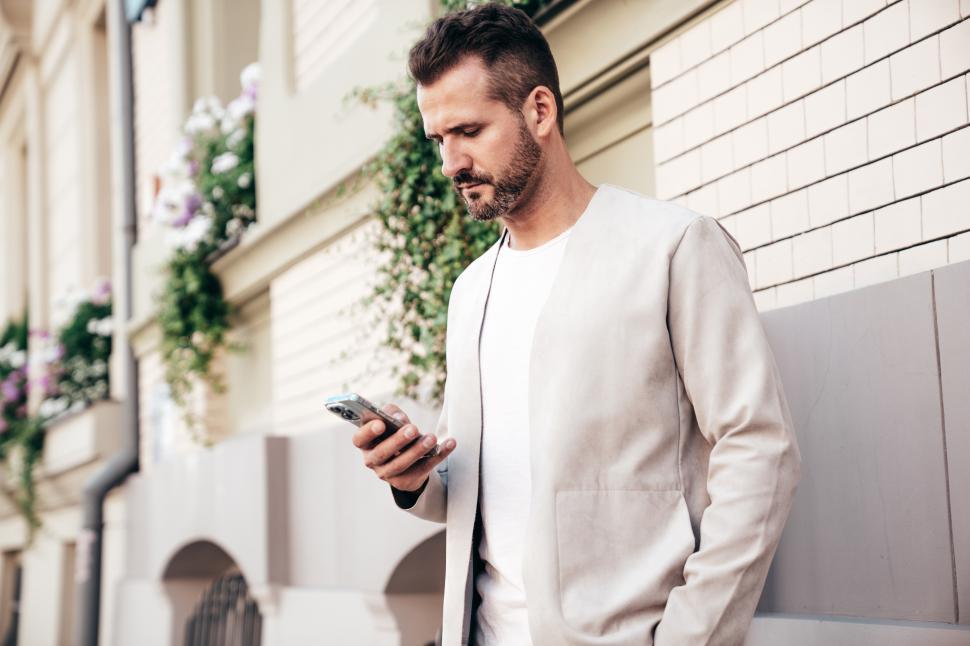 Free Image of Man in light jacket checking smartphone while standing against building adorned with flowers 