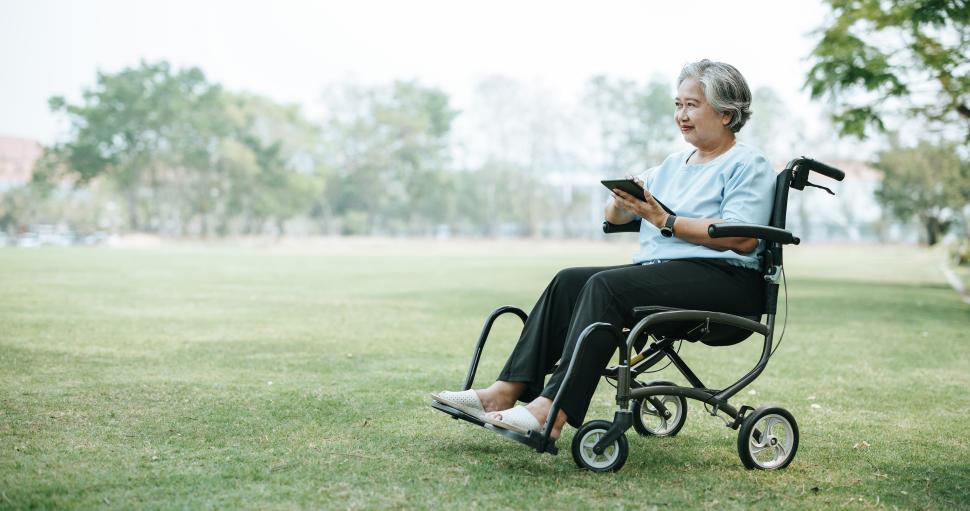 Free Image of Mature Asian woman on wheelchair use tablet at grass field 