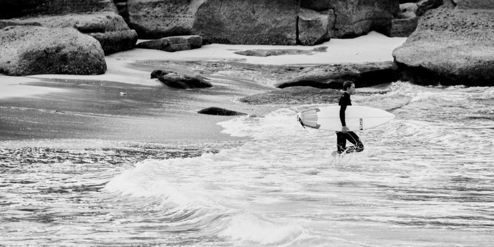 Free Image of Surfer carrying board walks into surf near large rocks. 