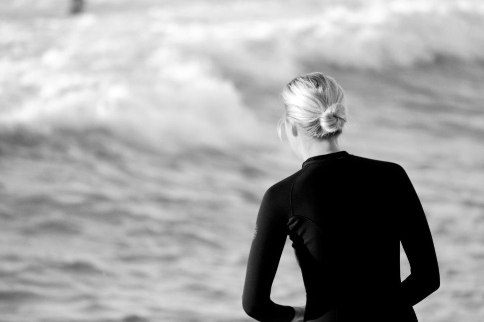 Free Image of A woman in a wetsuit looks at the ocean waves 